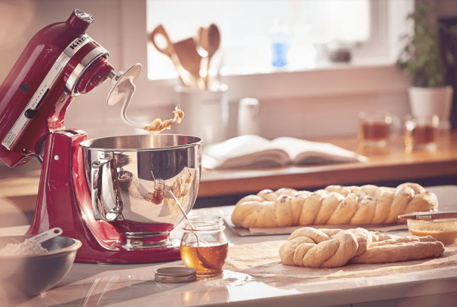 Batedor de Massas - Batedeira Artisan vermelha com gancho de massas em bancada de mandeira com massa de pães e jarra de mel.