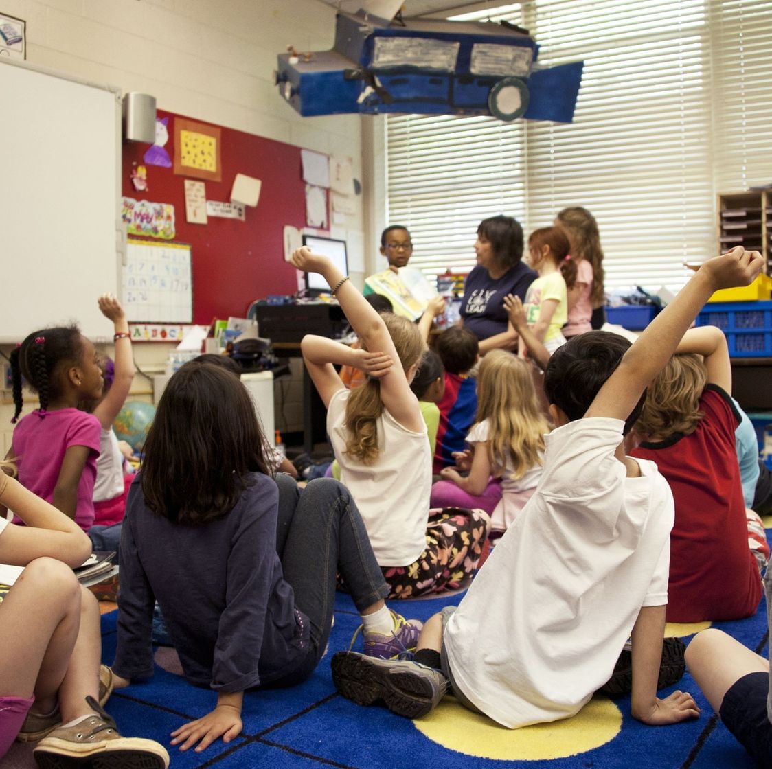 Alunos sentados em um tapete assistindo uma professora em sala de aula
