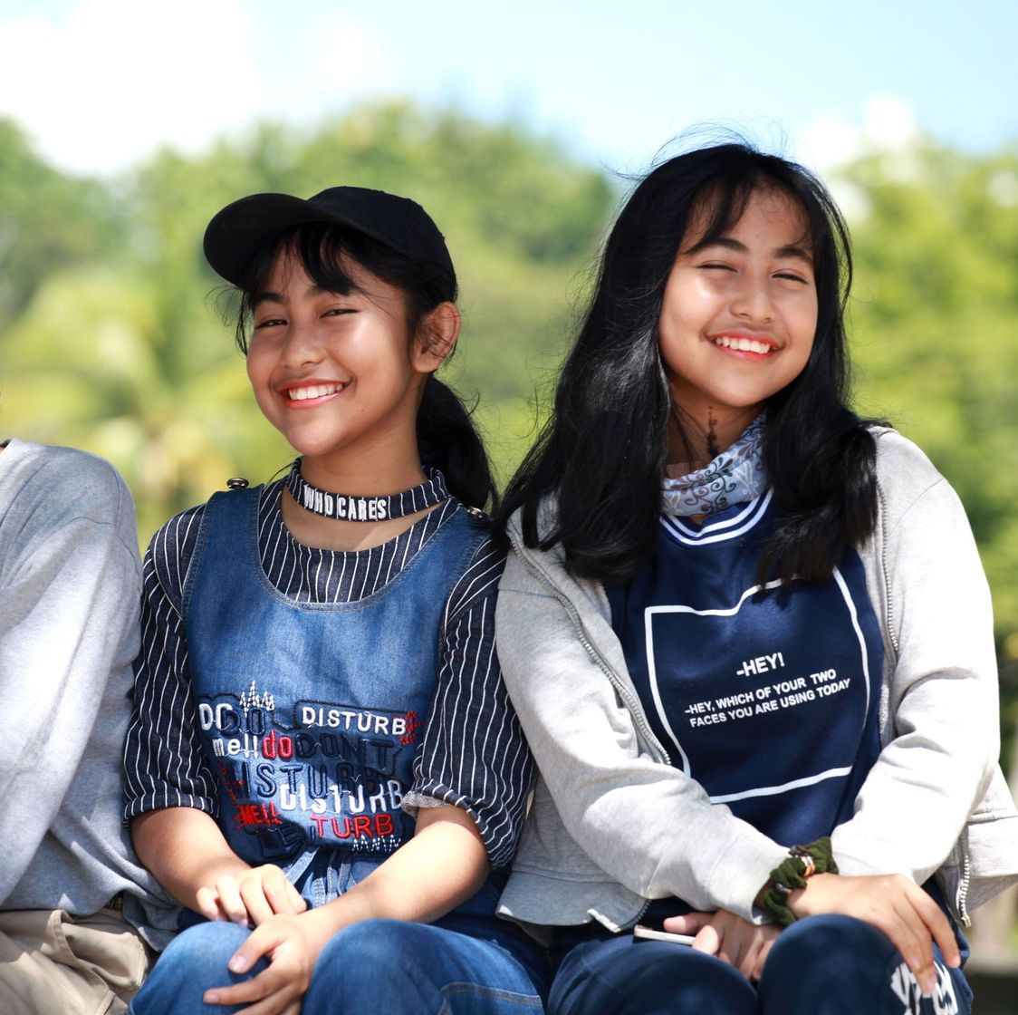 Duas garotas sentadas, sorrindo, ao fundo uma área arborizada