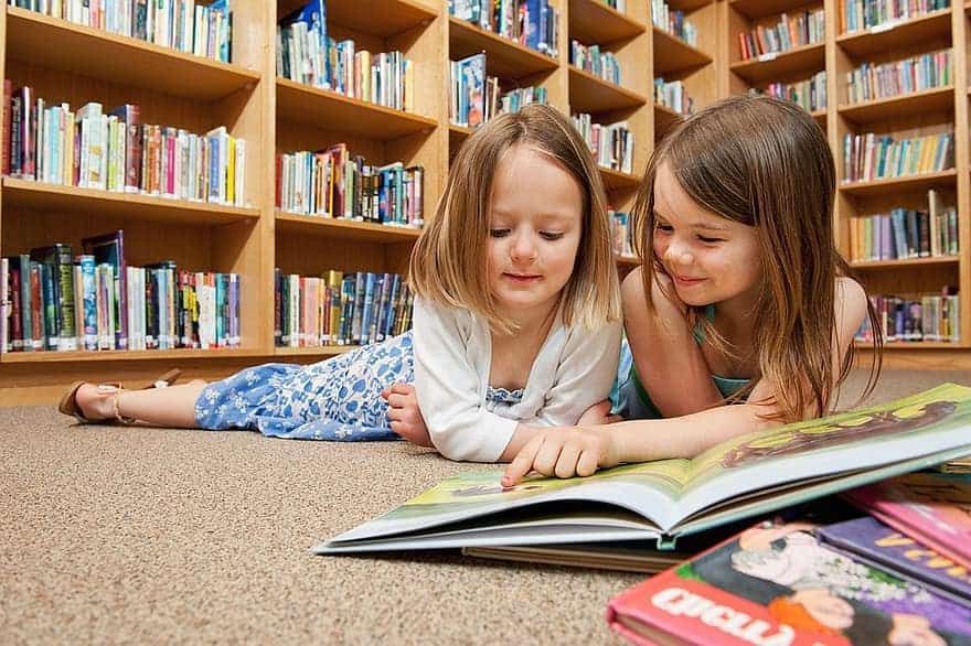 Duas crianças deitadas no chão de uma biblioteca lendo, mostrando a importância da leitura na educação infantil.