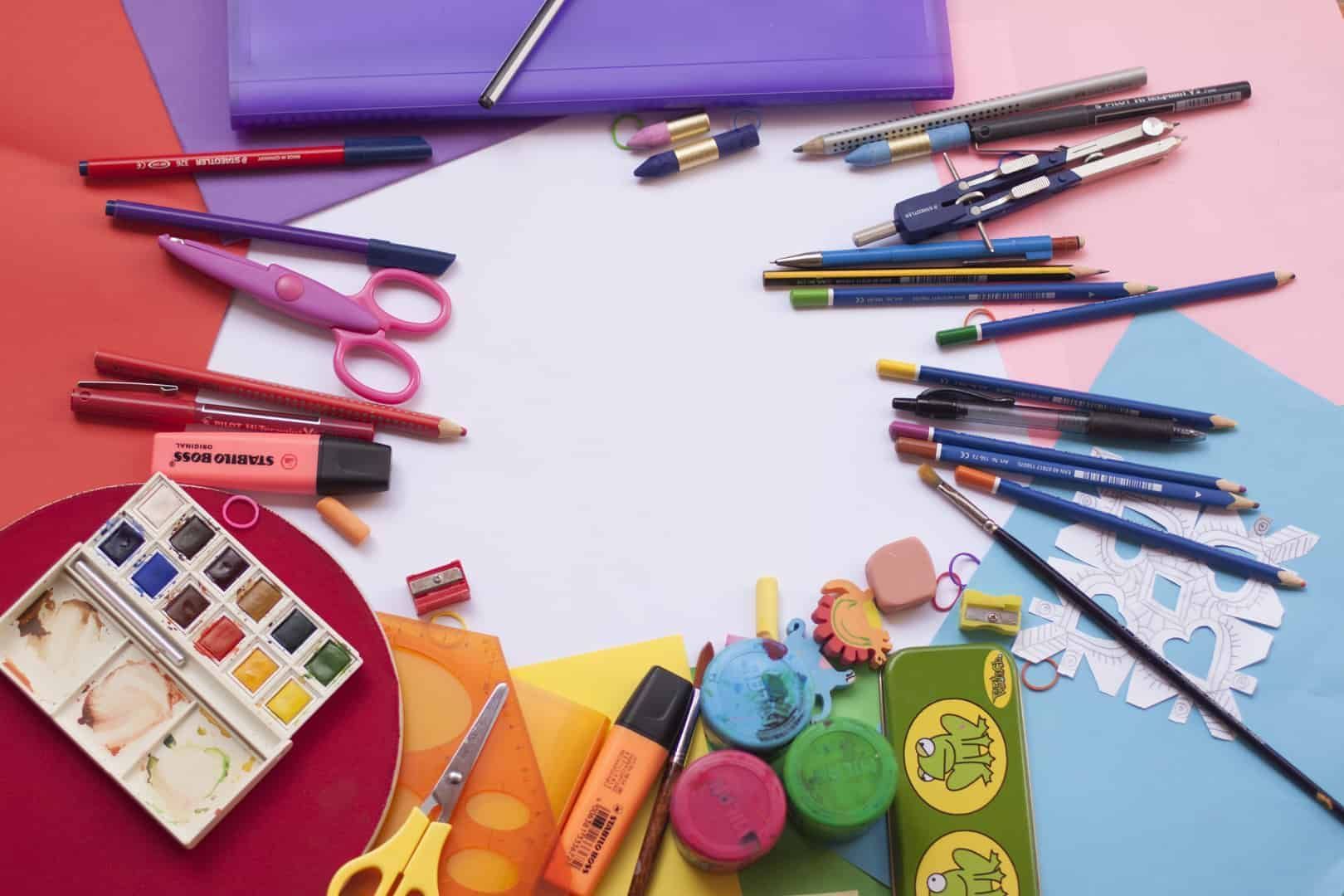 Foto mostra canetas, tesouras e outros materiais escolares dispostos em cima de uma mesa colorida. No novo normal, as papelarias online são uma ótima alternativa para as famílias que querem comprar sem sair de casa.