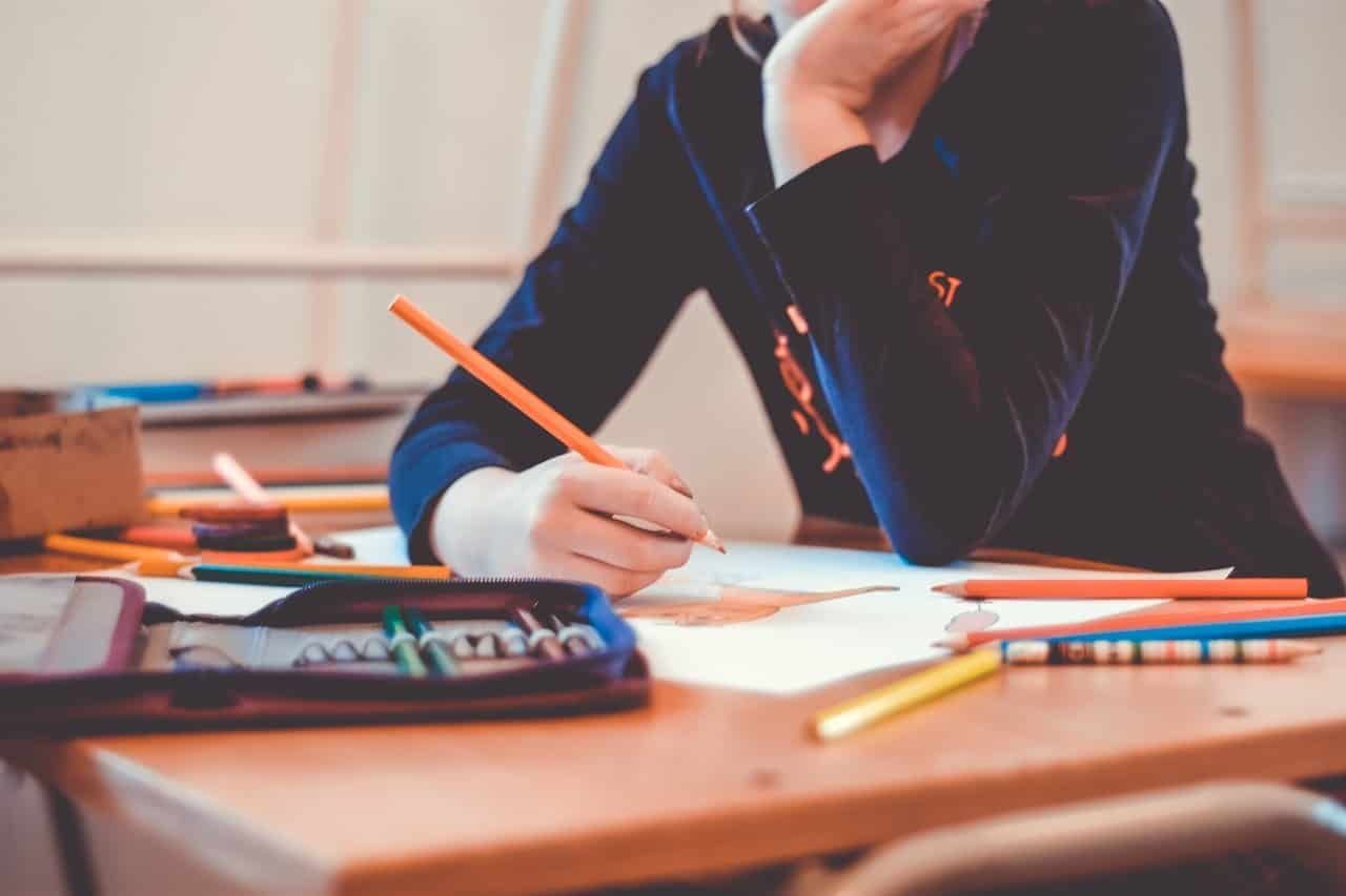 Menina adolescente segurando um lápis na mão, apoiando os braços em uma mesa cheia de material escolar. 