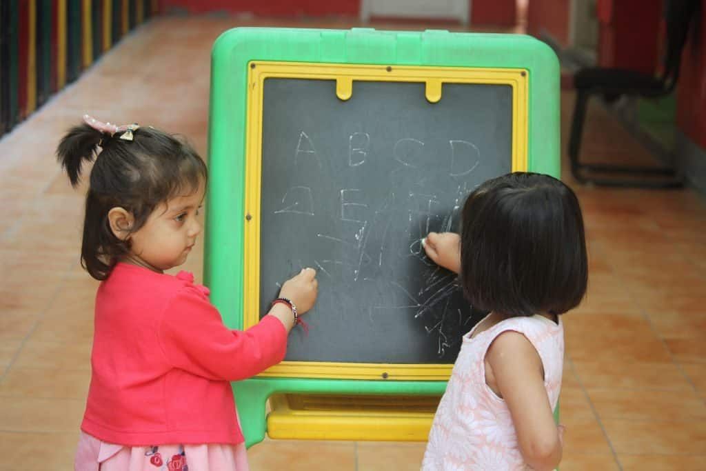 Duas meninas pequenas, em pé, escrevendo as letras do alfabeto em uma lousa que está apoiada no chão. Escrever e ler são uma das bases da alfabetização e do letramento.