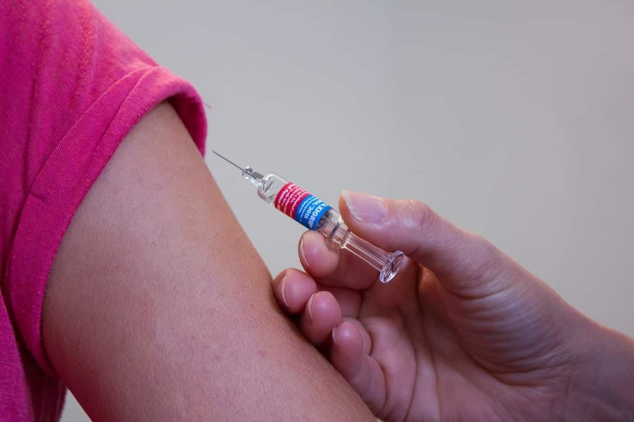 Uma criança de camiseta rosa está tomando vacina no braço. A vacina infantil é importantíssima na prevenção de doenças além de ser obrigatória a fim de matricular a criança em uma escola.