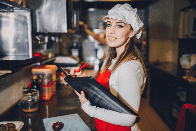 Mulher branca com avental e chapéu de cozinheiro escrevendo um menu dentro de uma cozinha industrial