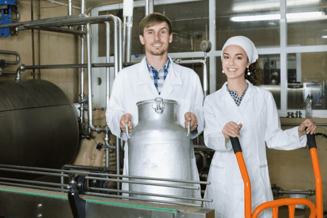 Um homem branco de avental e uma mulher branca de avental segurando silos de leite em uma fábrica produtora de leite animal