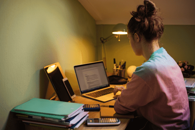 Mulher branca ruiva estudando pelo notebook em uma escrivaninha com vários livros e calculadora próximo na sua casa