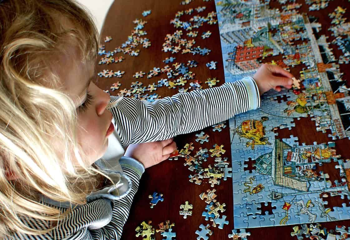 Menina loira montando um quebra-cabeça na mesa. O jogo é uma ótima ferramenta para o desenvolvimento de habilidades cognitivas, motoras e sociais nas crianças.