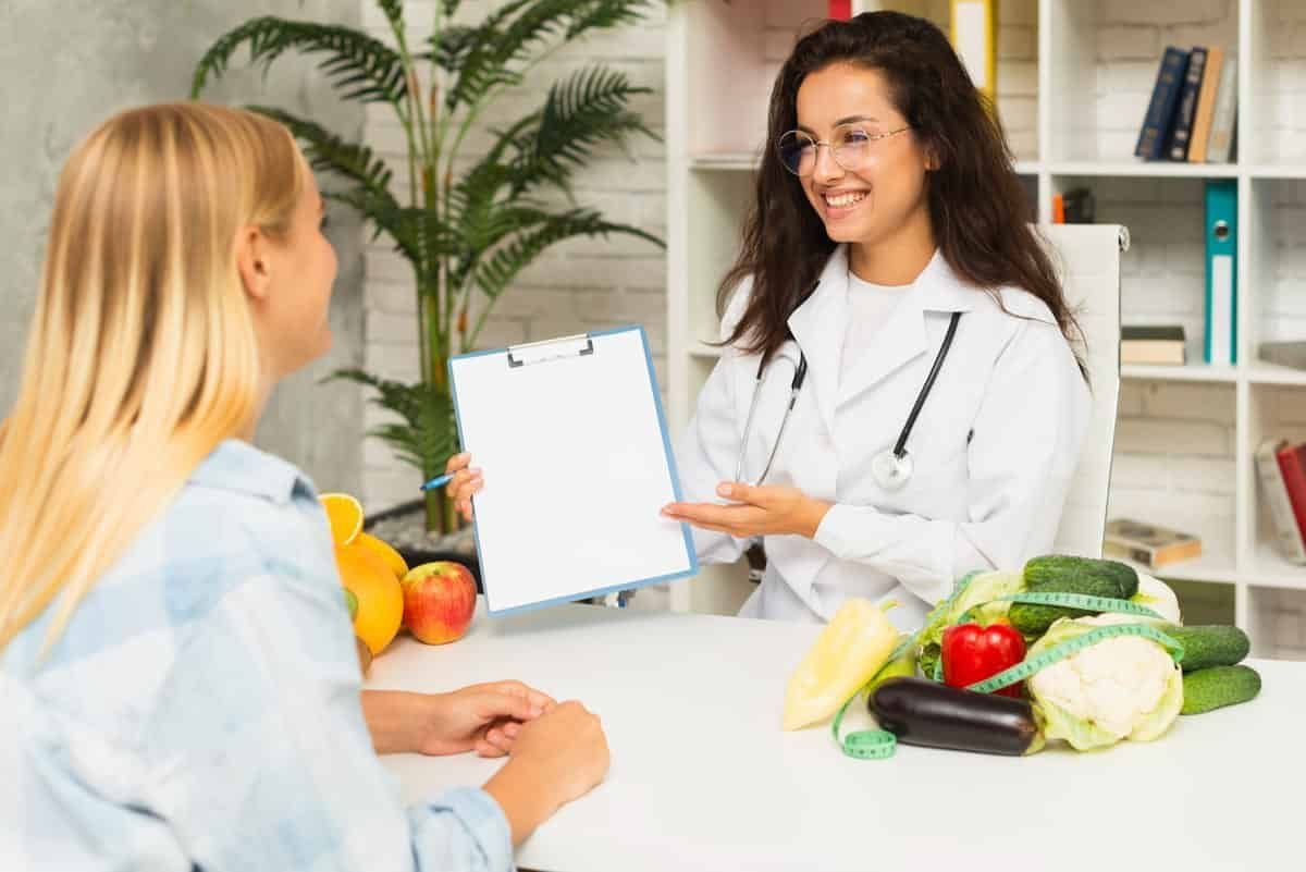 Nutricionista formada em Nutrição à distância (EaD) mostra prancheta a paciente que está sentada em frente a uma mesa repleta de alimentos saudáveis, no consultório