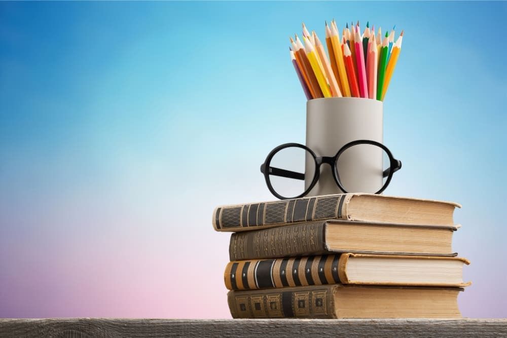 Uma pilha de livros, um conjunto de lápis e um óculos integram a imagem. 