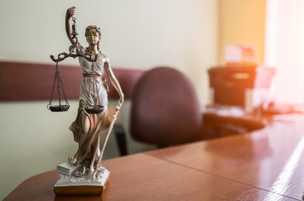 Estatueta do símbolo da Justiça possui cor similar ao tom de cobre e está localizado em cima de uma mesa de madeira.