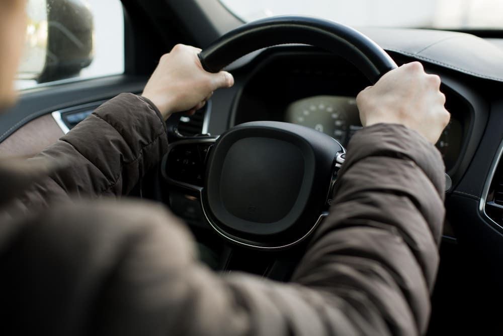 A imagem mostra uma pessoa dirigindo carro, sentada no banco de motoristas e com as duas mãos no volante.
