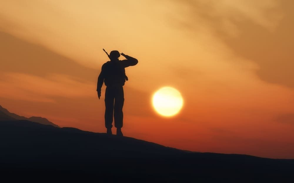 Soldado, parado em cima de terreno ondulado, observa por do sol. 