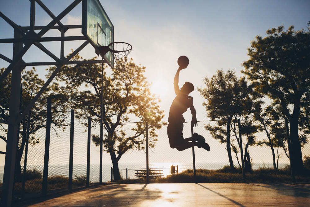 Homem segura bola de basquete em suas mãos, durante salto, em quadra esportiva. O movimento realizado pelo atleta é conhecido como enterrada no basquetebol.