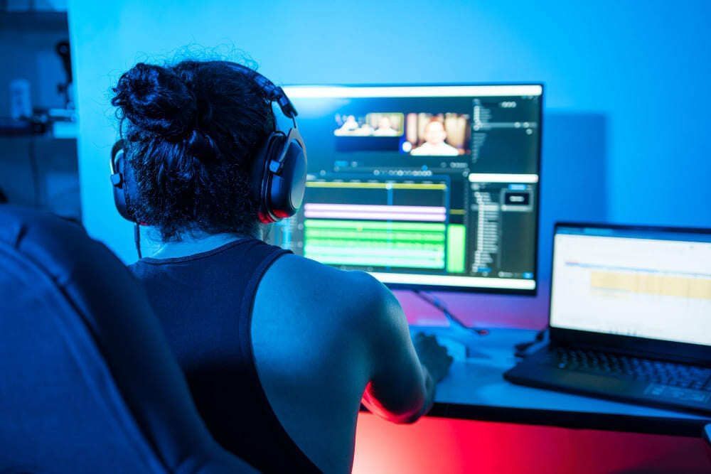 Pessoa está em frente ao computador com um editor de áudio aberto. O local em que a pessoa trabalha possui uma iluminação azulada e o indivíduo está de costas no plano da imagem,