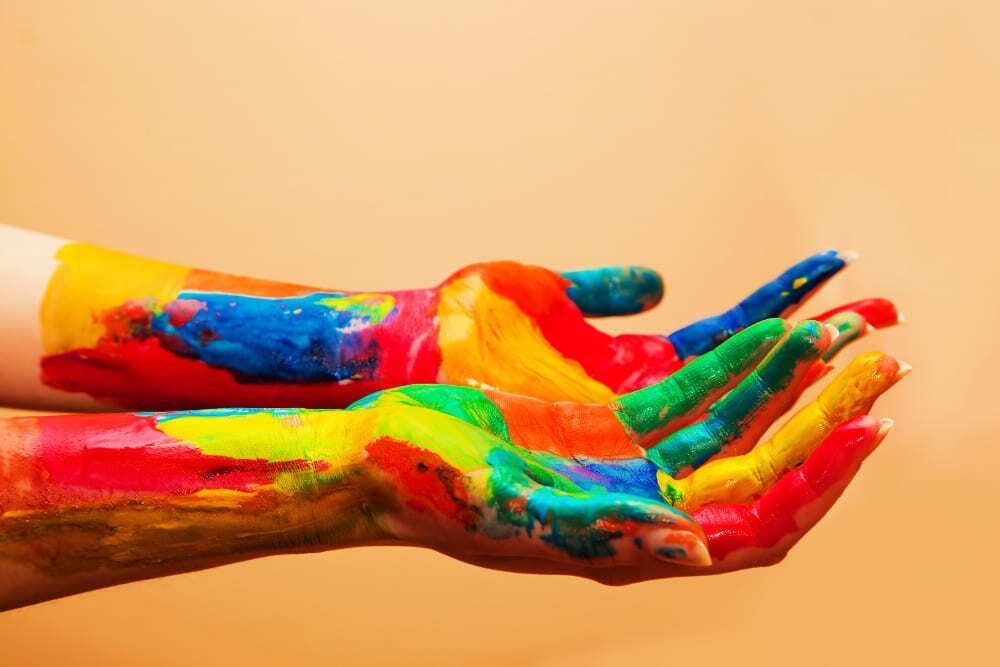 A imagem retrata duas mãos cobertas de tintas. São diversas cores nas duas mãos, que estão abertas na imagem: vermelho, amarelo, laranja, verde, azul e outras.