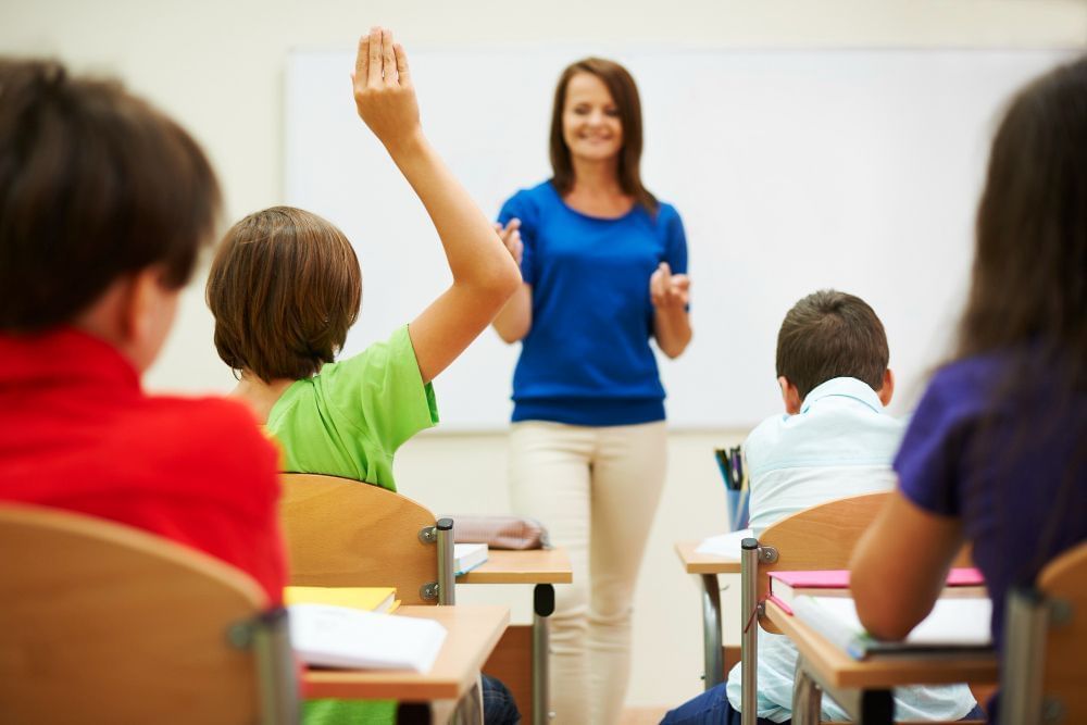 Imagem retrata sala de aula e aluno levanta a mão para questionar professora