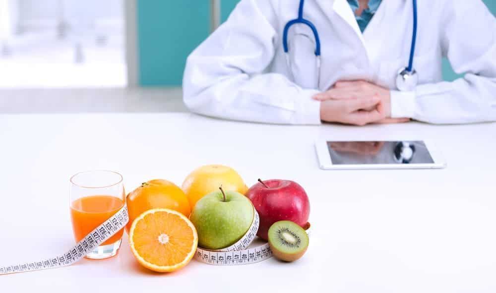 Imagem é composta por: Nutricionista sentada em cadeira e, em sua mesa, estão frutas, um copo de suco e uma fita métrica.