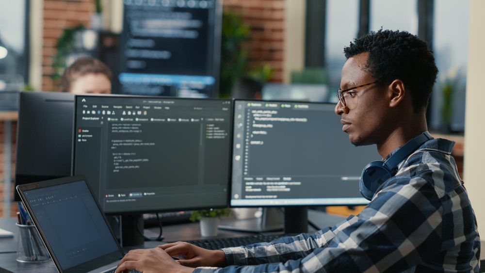 imagem retrata profissional trabalha com computador e dois monitores em ilha de produção da área de tecnologia, software e dados. 