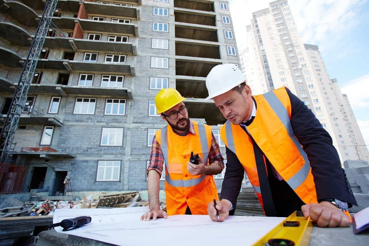 Dois homens engenheiros civis com equipamentos de segurança, olhando a planta de um edifício. Ao fundo a obra deste edifício.