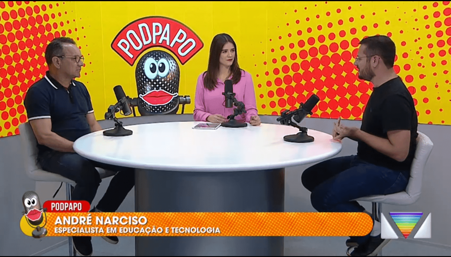 Dois homens e uma mulher sentados em volta de uma mesa redonda, com microfones para podcast, em um fundo amarelo com bolinhas laranjas, onde está escrito "PodPapo"