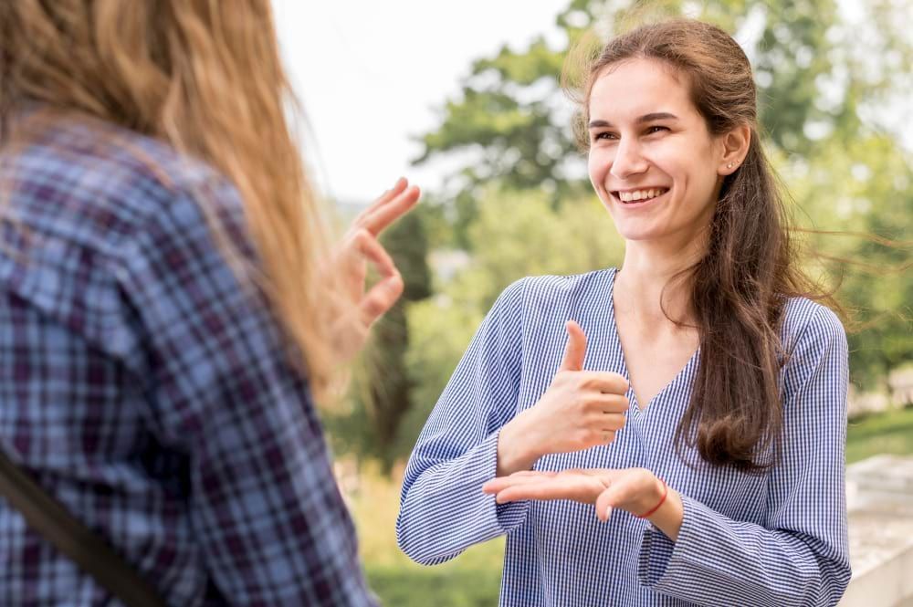 Na imagem, duas mulheres brancas de cabelos castanhos se comunicam em línguas de sinais, uma de frente para a outra. Uma delas está de costas e a outra sorri enquanto reproduz os gestuais da língua de sinais.