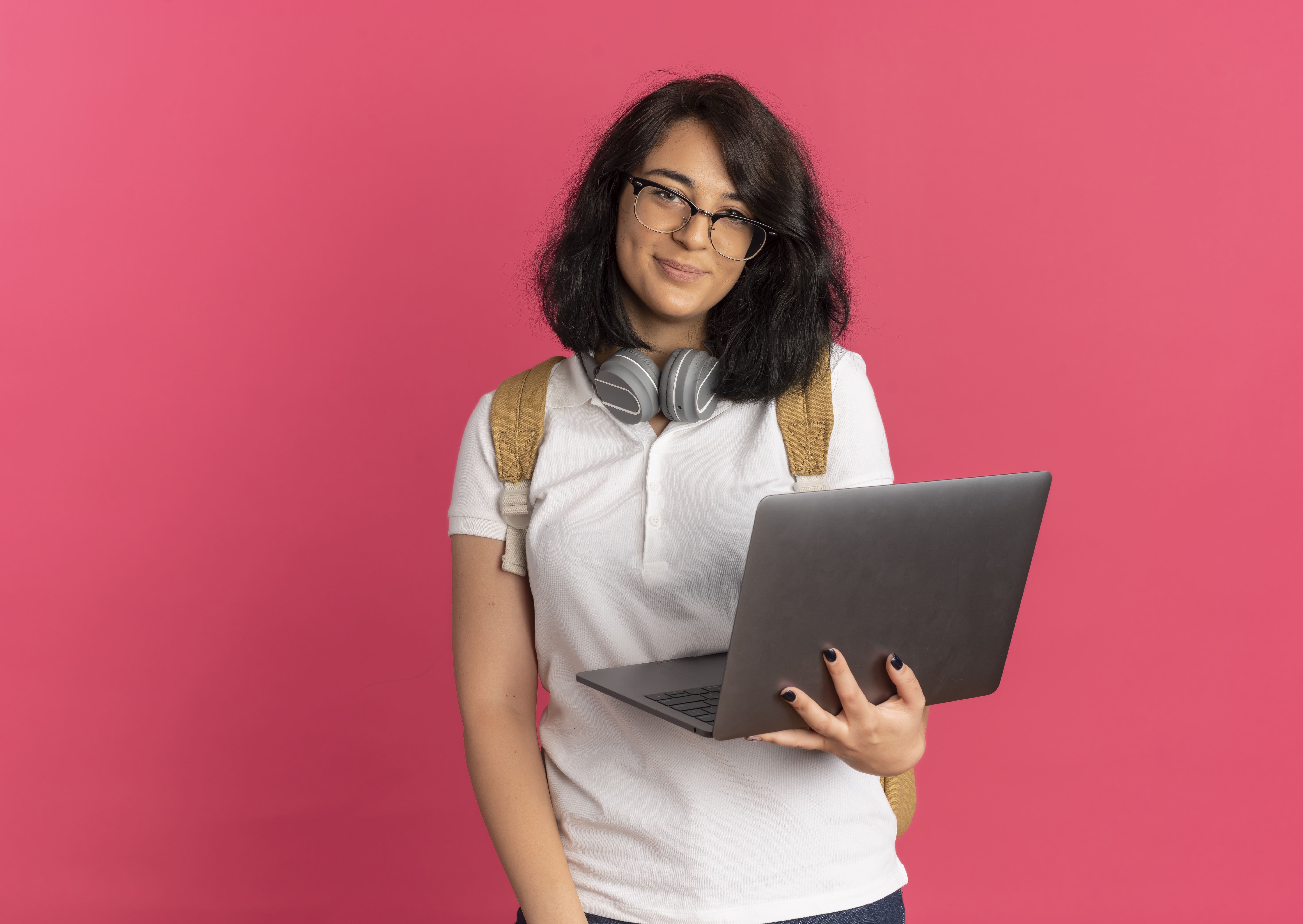 mulher de cabelo preto com óculos, fones pendurados no pescoço, mochila nas costas segurando um notebook. Ela usa camiseta branca e o fundo da imagem é rosa.