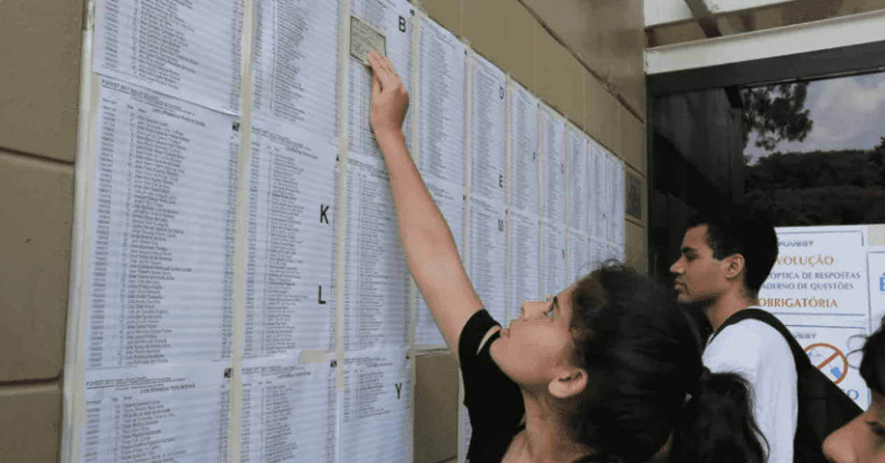 Jovem mulher olhando para a lista de candidatos aprovados em folhas brancas; Fuvest 2024: aprovados em lista de espera são divulgados