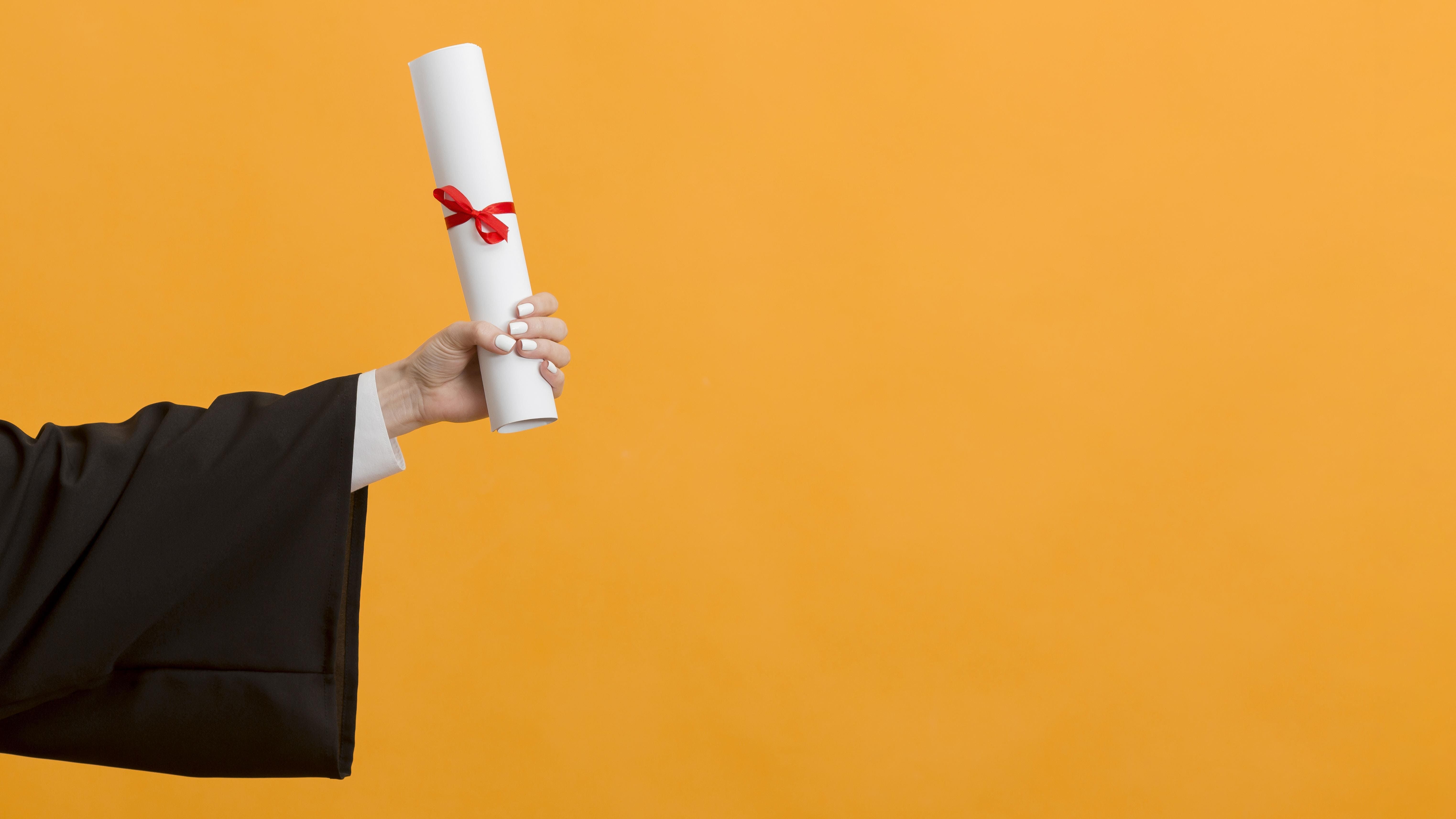 Braço de pessoa vestida com beca de formatura segurando um diploma em fundo laranja