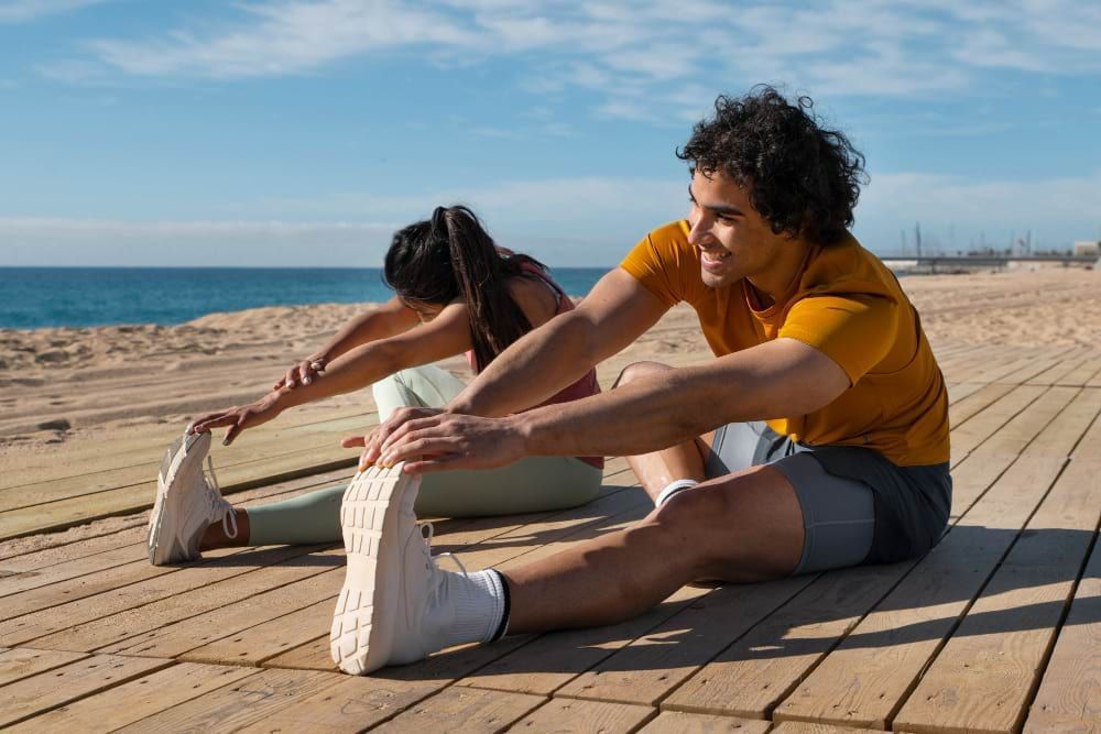 Um homem e uma mulher estão se alongando em um deck em frente a praia. Estão alegres e se preparando para se exercitar.
