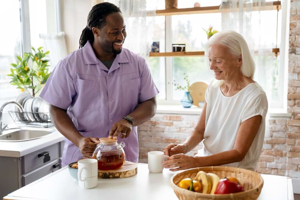 Um homem e uma senhora idosa estão em uma cozinha preparando um chá. Ele parece ser um profissional da saúde, cuidados e auxiliar de idosos, e ela sua paciente. Ambos estão felizes e tranquilos.