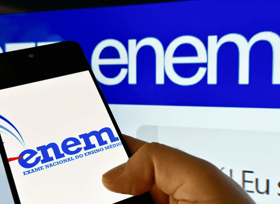 Mão segurando um celular que mostra o logo do Enem, ao fundo, uma tela de computador com o logo da prova; saiba como fazer a solicitação de isenção do Enem