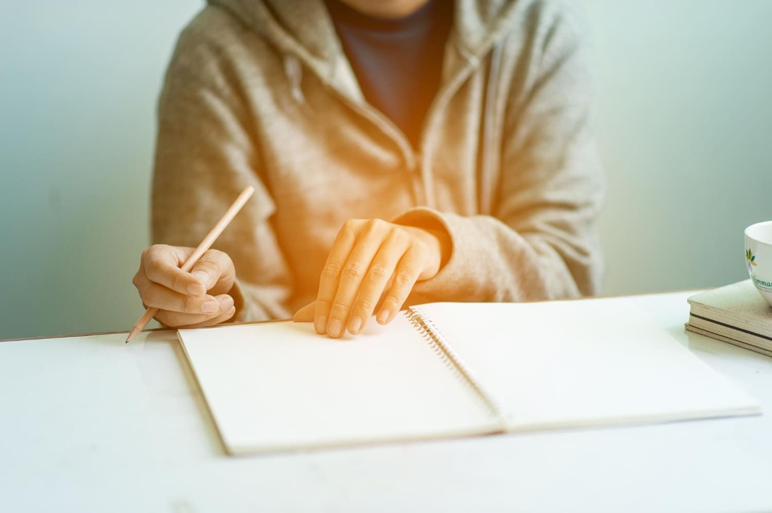 Mãos de pessoa segurando um lápis e folheando um caderno sobre uma mesa tentando fazer uma redação de 20 linhas