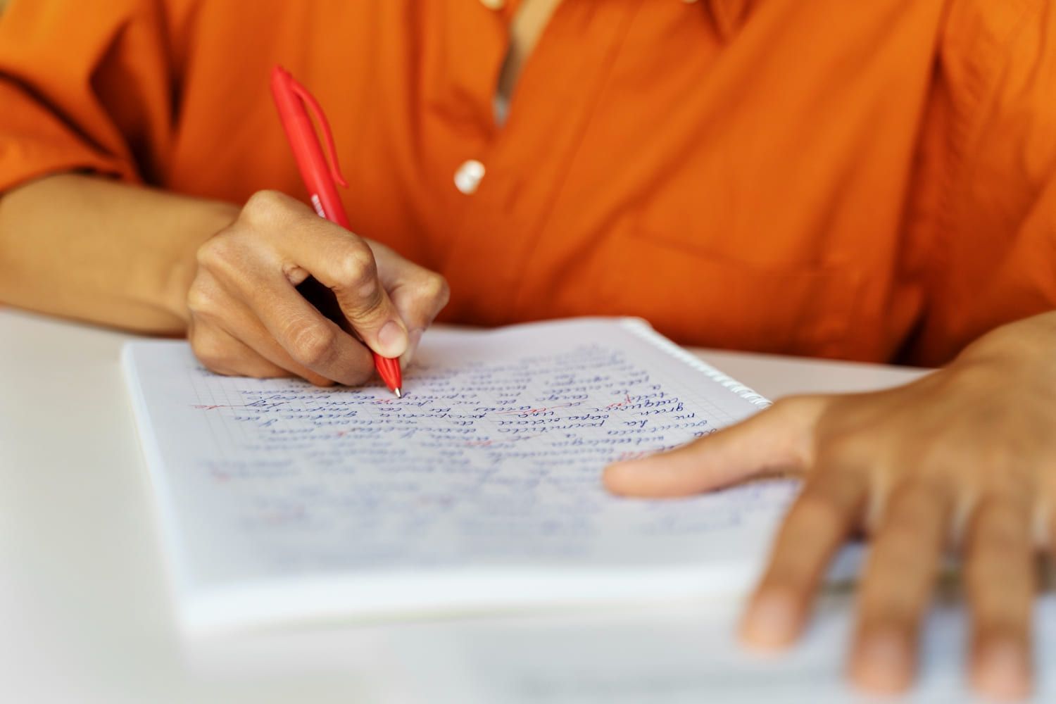 Pessoa sentada com mãos em cima da mesa escrevendo redação concisa e de 20 linhas em folha sulfite