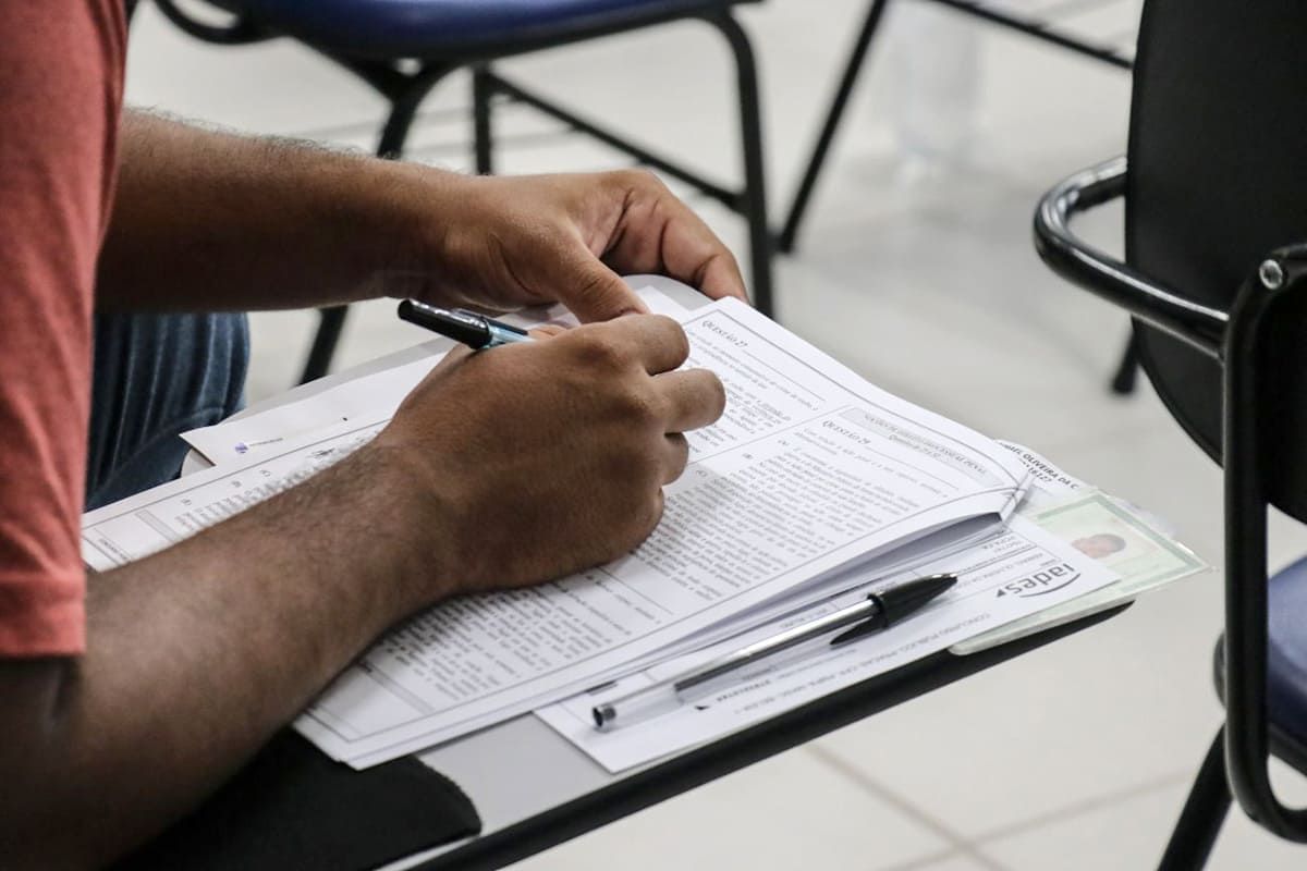 Pessoa sentada em uma cadeira escrevendo com mão em cima da prova; saiba como ver o local de prova do CNU
