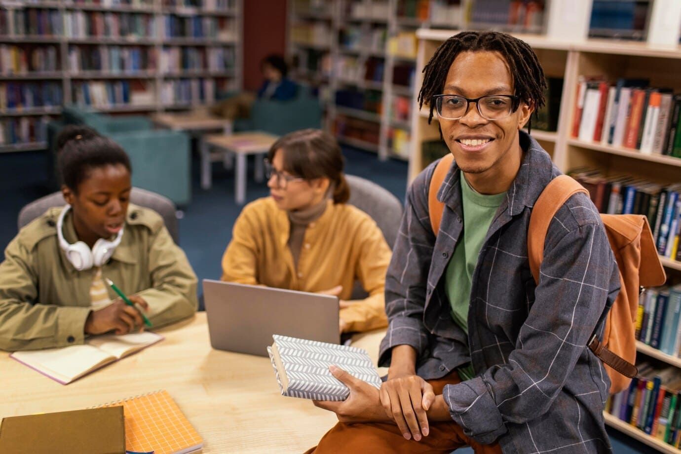 3 estudantes de um curso técnico numa biblioteca, com duas mulheres sentadas, uma branca e outra negra em segundo plano, enquanto mais a frente temos um homem negro com mochila sentado em cima da mesa