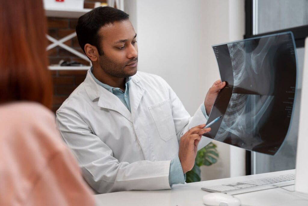 técnico em radiologia, homem negro, com camisa social azul e jaleco branco durante o atendimento mostrando ao paciente um exame de raio-x.