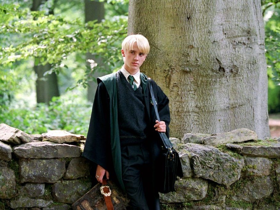 Draco Malfoy apoiado num muro de pedras com fundo que parece uma floresta, verde roupa da sonserina