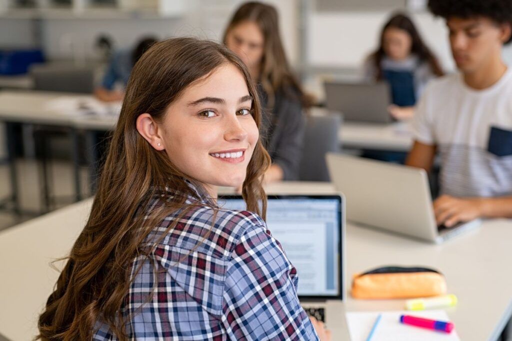 Estudante, mulher, sorrindo com cabelo castanho e camiseta quadriculada estudando numa sala de aula