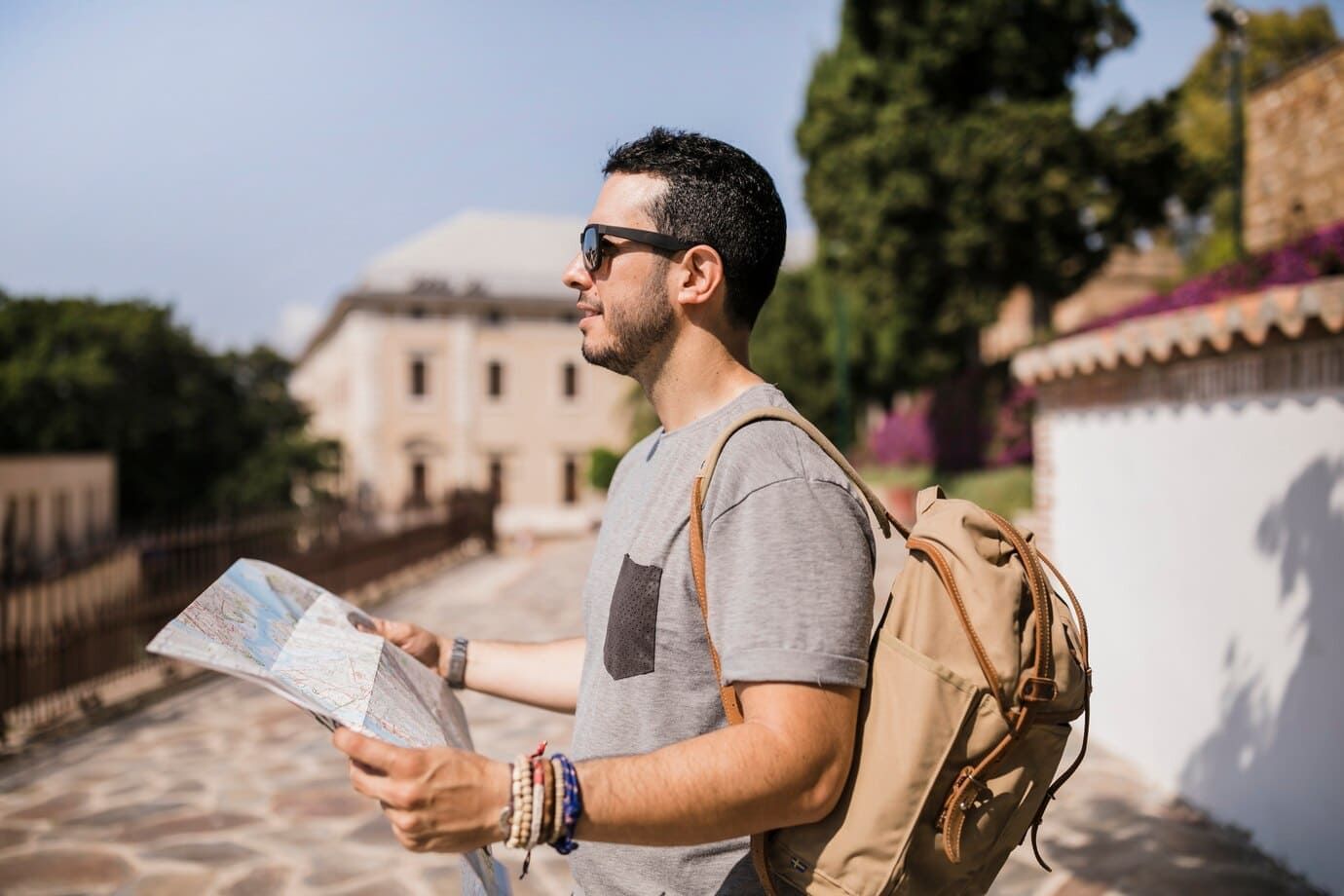 Guia de Turismo, homem, branco, com óculos de sol, de lado para a imagem, com camiseta cinza e mochila bege segurando um mapa.