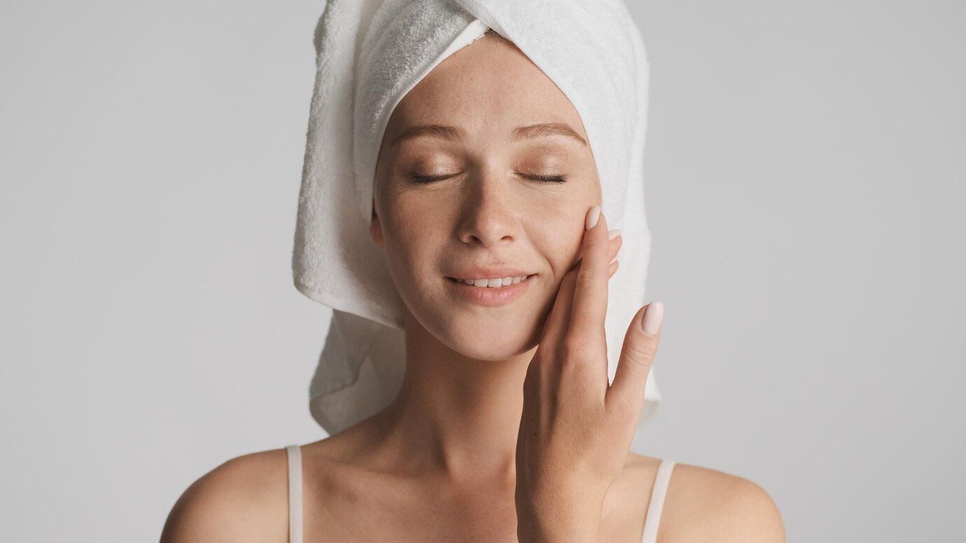 Mulher branca com toalha presa na cabeça numa clínica de estética, passando as mãos na cara após tratamento com técnica em estética.