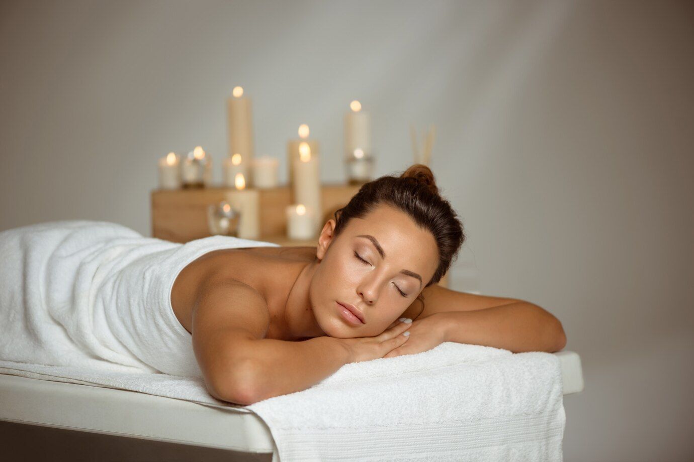 Mulher branca, com cabelo castanho recebendo uma massagem, deitada, com olhos fechados, relaxando, com toalhas envolvendo seu corpo.