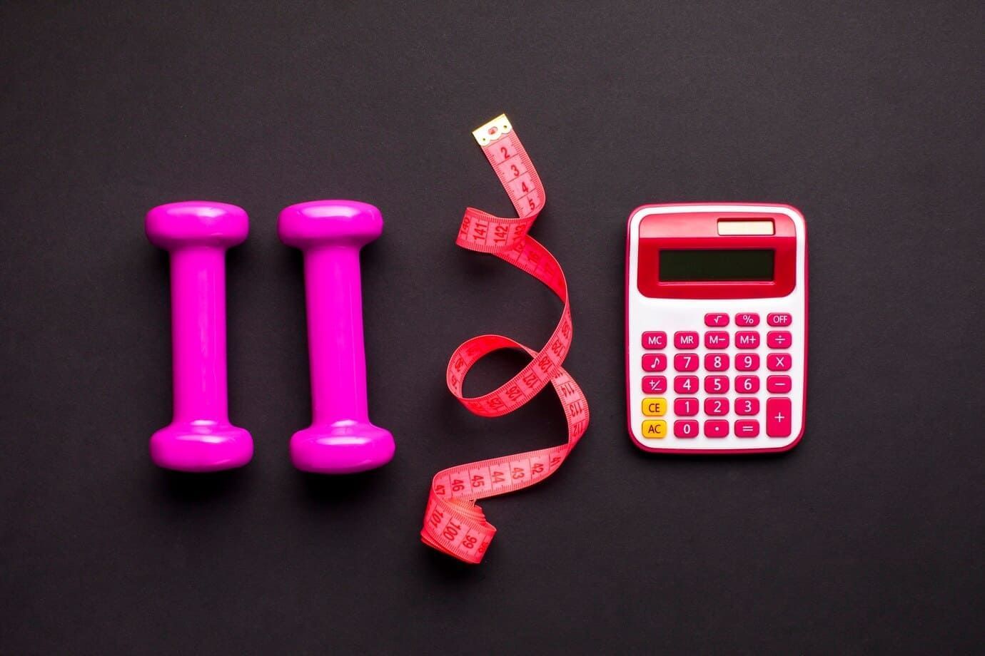Dois halteres rosa, uma fita métrica vermelha e uma calculadora branca e vermelha colocadas num chão preto.