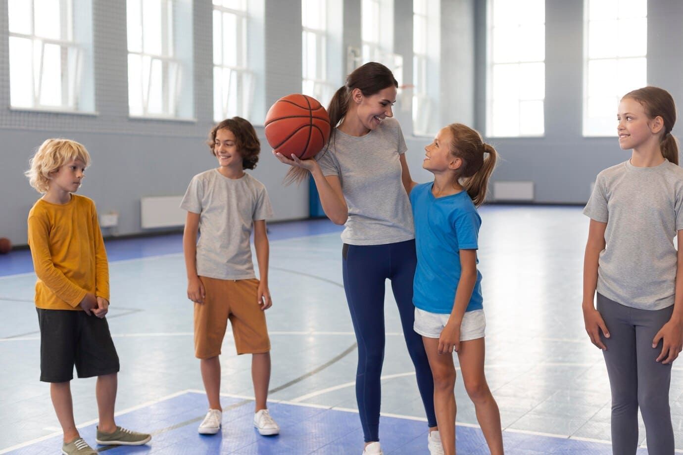 Professora de educação física, dando aula, mulher branca, com cabelo castanho longo, camiseta cinza e calça legging azul, segurando uma bola de basquete, dando aula para 4 alunos, duas meninas e dois meninos.