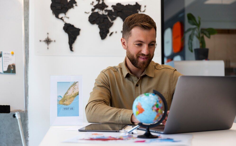 Homem, branco, com cabelo castanho e camiseta marrom clara, agente de viagens, trabalhando em seu computador. Parece trabalhar num escritório, com um mapa ao fundo e apoiado na mesa há um tablet e também um globo terrestre.