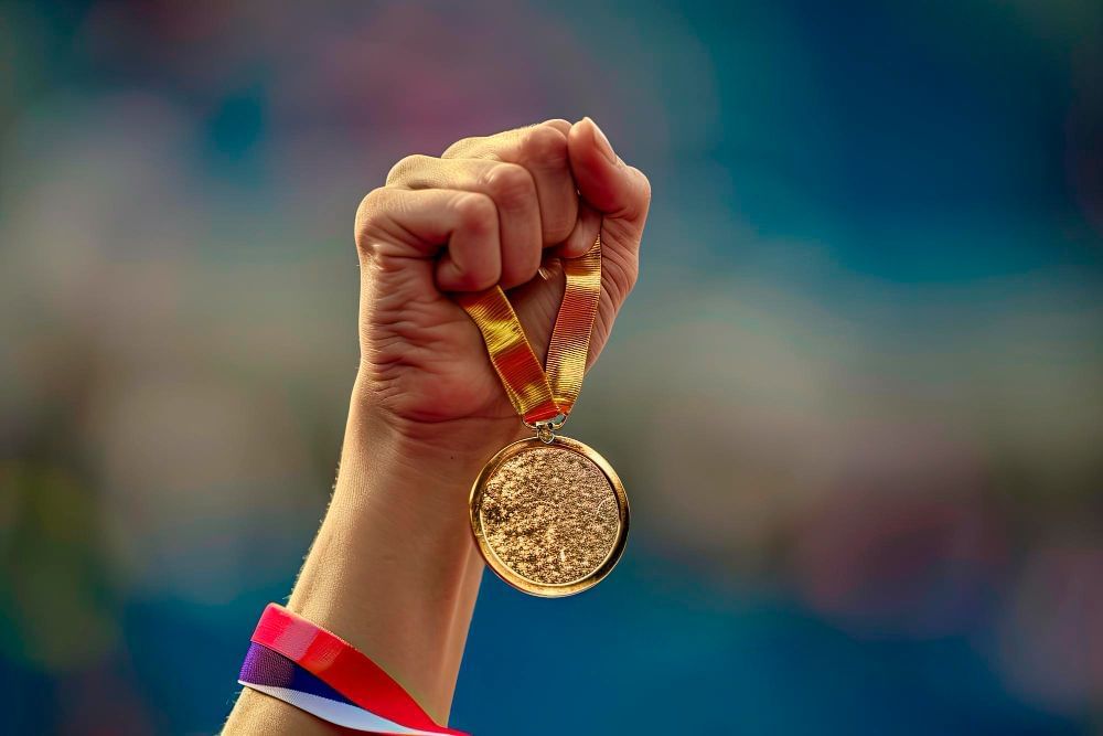 A mão de uma atleta olímpica é vista erguida para cima segurando uma medalha de ouro.