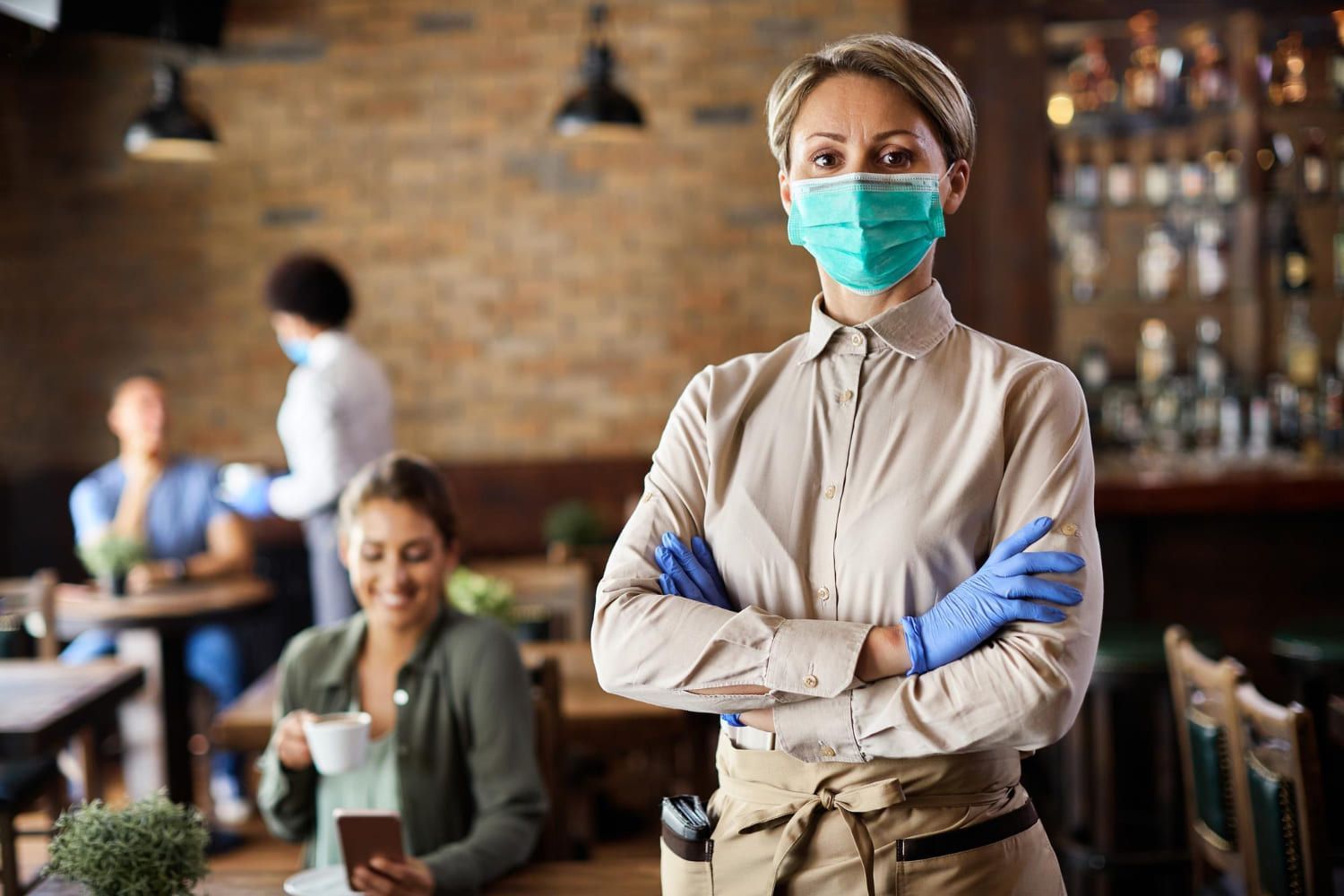 Mulher profissional Técnica especializada em Higiene Ocupacional de mãos cruzadas em frente a um restaurante, vestindo luvas e máscara