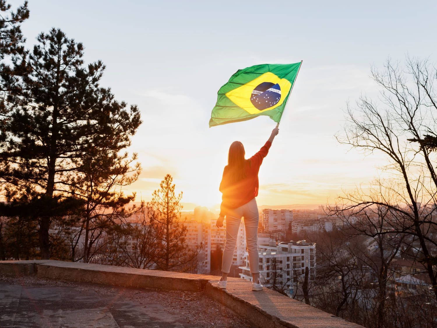 Mulher em um lugar alto, olhando para o sol se pondo em cima da cidade, segurando no alto a bandeira do Brasil; entenda o que se comemora no 7 de setembro e qual a importância da Independência do Brasil