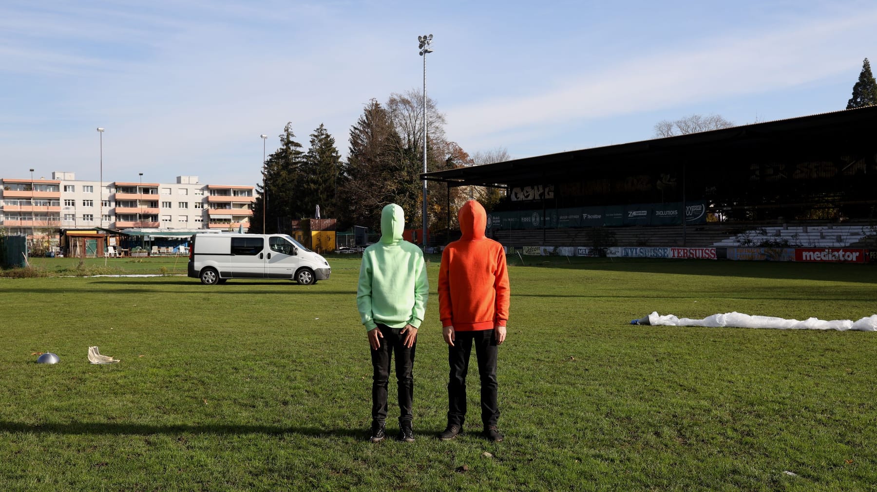 Auf einer grossen Wiese, im Hintergrund sind Gebäude und ein Auto, stehen zwei Personen, die Hoodies in mintgrün und rot tragen, jedoch so herum, dass die Kapuzen ihre Gesichter verdecken.