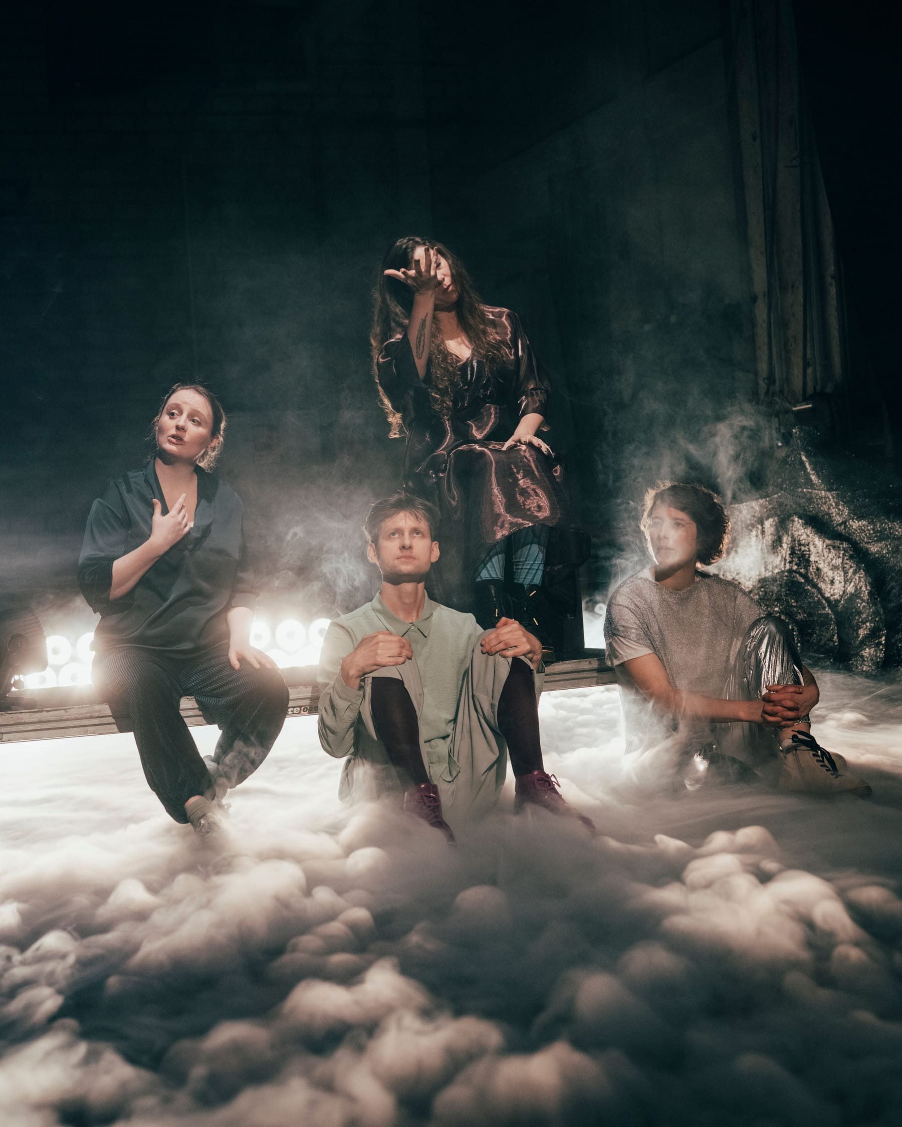 Vier Personen auf einer Bühne mit viel Rauch am Boden. Im Vordergrund sitzen drei Personen und eine Person steht im Hintergrund. Alle haben den Blick nach oben gerichtet.